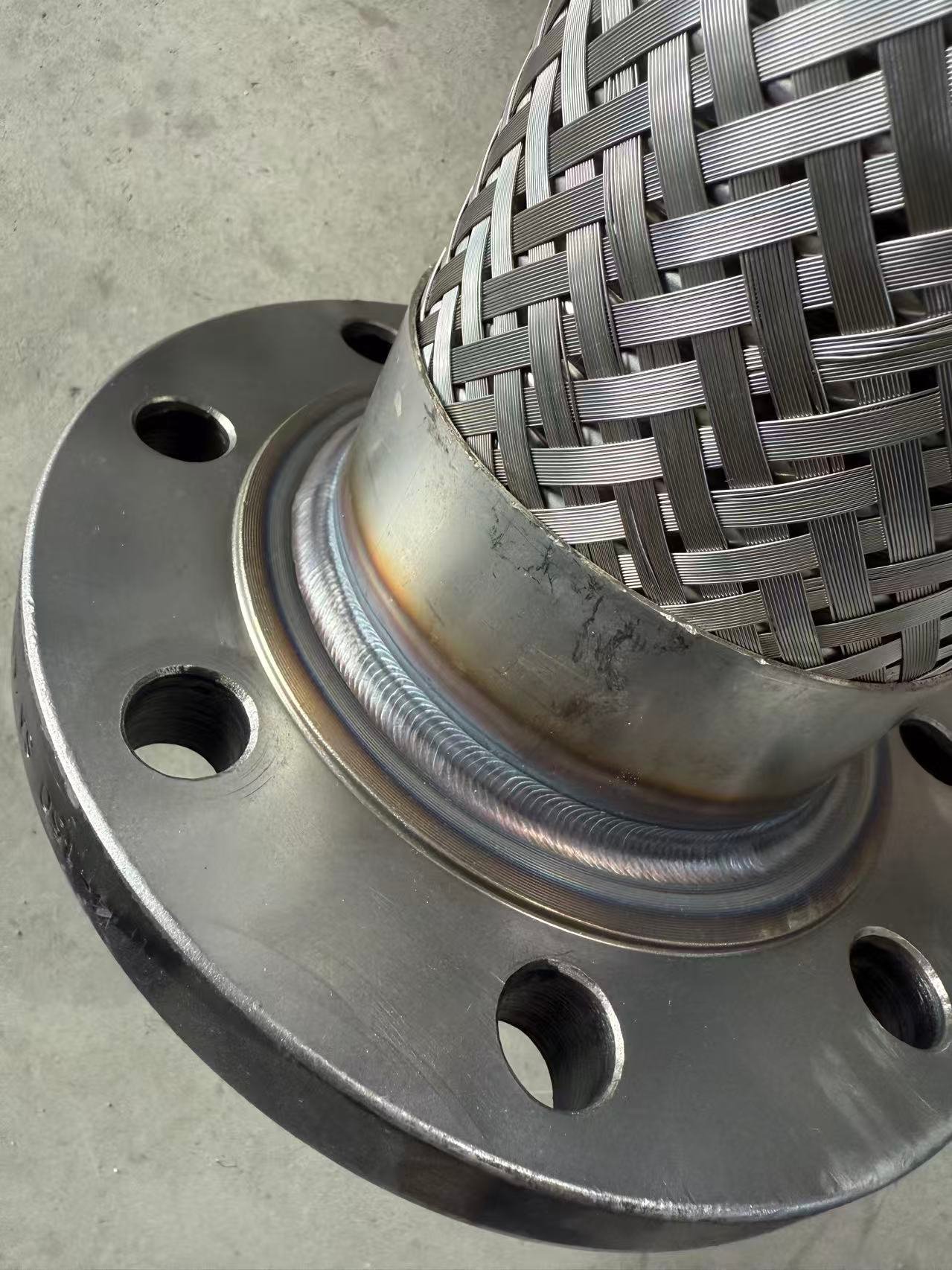 TIG-welded braided metal hose connected to a stainless steel flange with a uniform circumferential weld seam.
